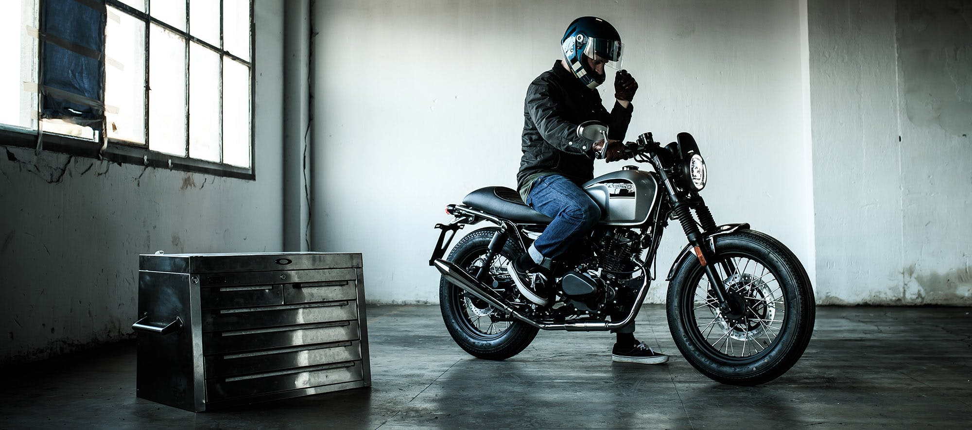 Rider sitting on his parked Brixton Felsberg 250 in Timberwolf Grey in an empty garage