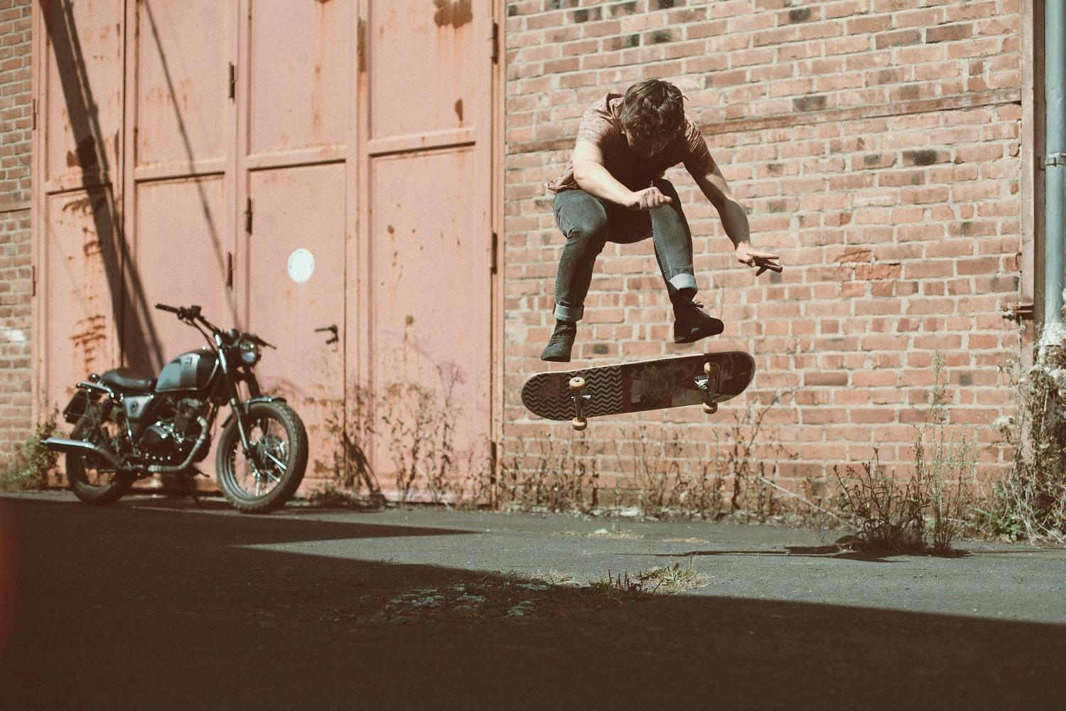 Rider of the Month Sidney doing a kick flip on a skateboard with his Brixton Cromwell 125 in Timberwolf Grey parked in the back