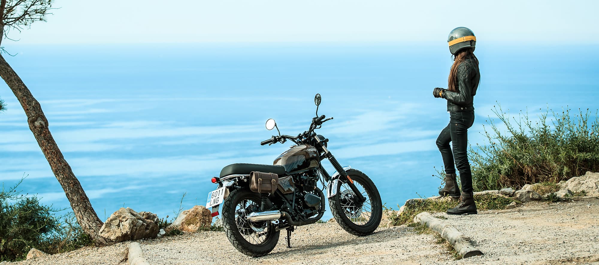 Female rider standing next to her parked Brixton Cromwell 125 in Charly Brown looking out over the cliff onto the ocean