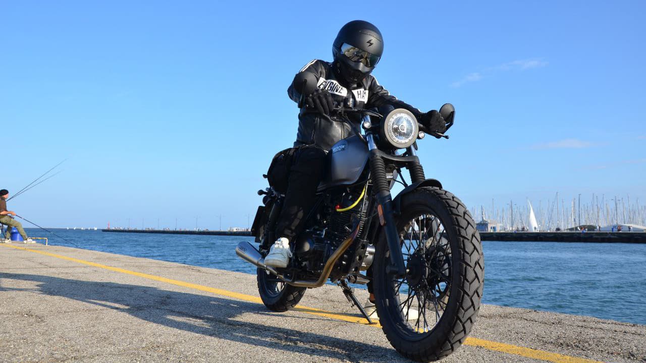 Rider of the Month Chris sitting on his Brixton Felsberg 125 in Timbewolf Grey at the pier with sailing boats and water in the back