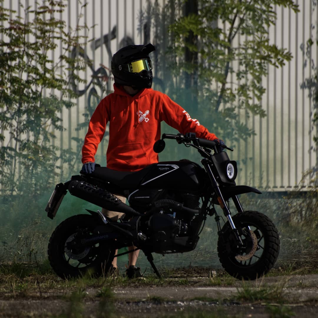 Rider of the Month Marco wearing a red hoodie, a motorcycle helmet and googles, standing next to his Brixton Crossfire 125 XS in Backstage Black