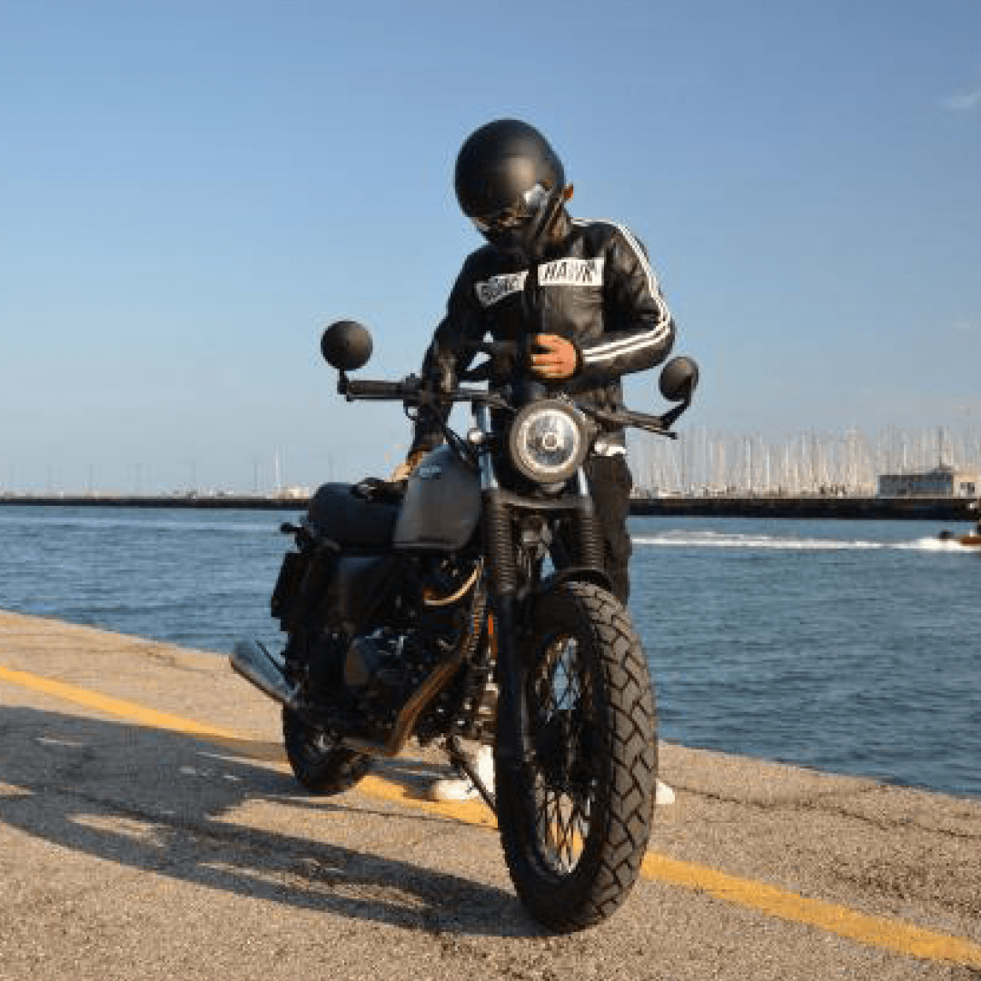 Rider of the Month Chris sitting on his Brixton Felsberg 125 in Timbewolf Grey at the pier with sailing boats and water in the back