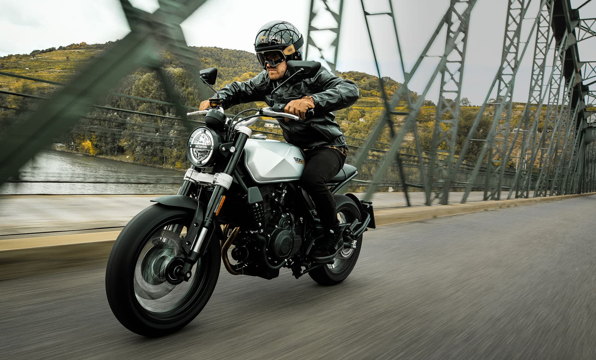 A rider wearing a leather jacket and gloves as well as a classic motorcycle helmet and googles riding a Brixton Crossfire 500 in Bullet Silver over a bridge above a river in the countryside