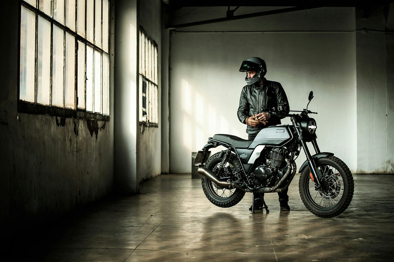 Rider in a full helmet, leather jacket and leather gloves standing behind his Brixton Motorcycles Felsberg 250 in Timberwolf Grey that's parked in an empty industrial warehouse