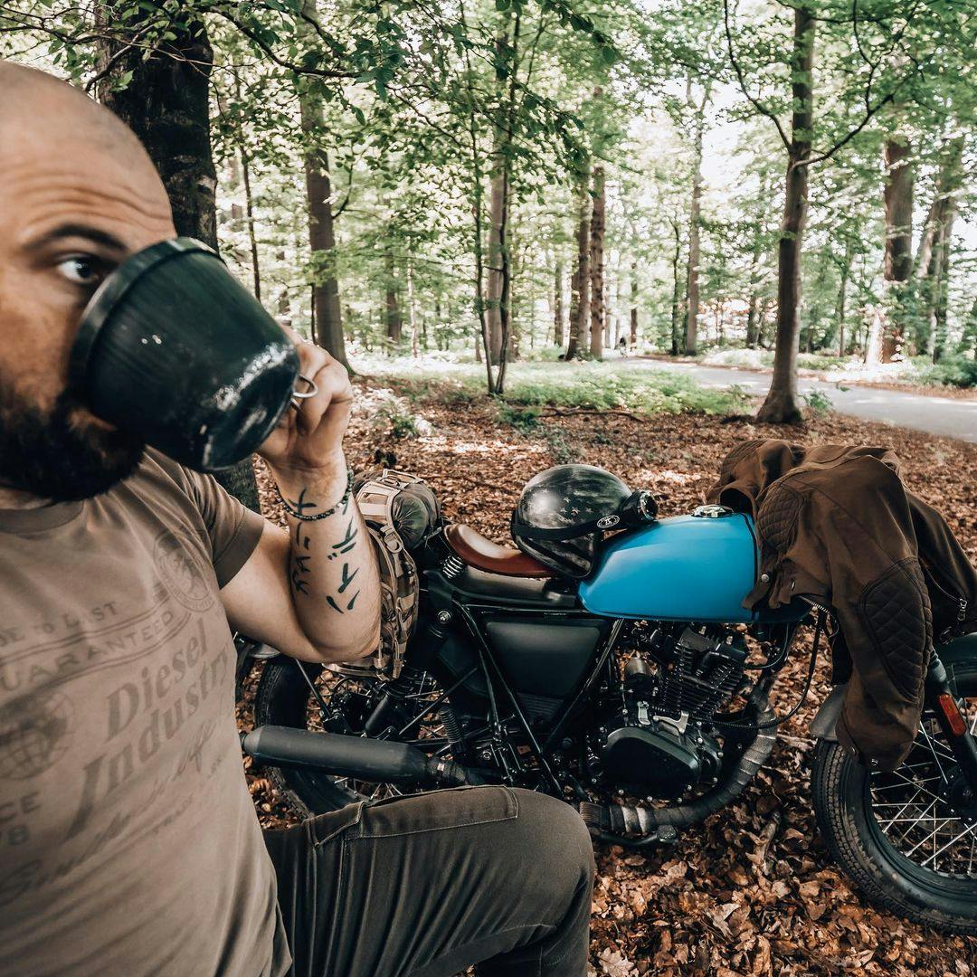 Lifestyle photo of Rider of the Month Kev drinking out of a cup while his Brixton Rayburn 125 in Royal Blue Matt / Horizon Matt is parked next to him in the forest