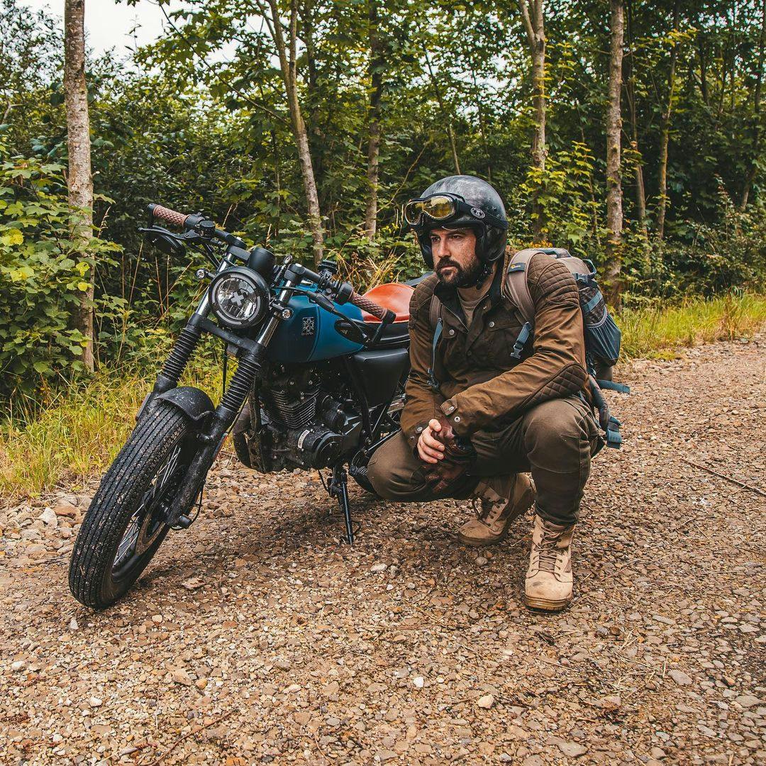 Rider of the Month Kev grouching next to his Brixton Rayburn 125 in Royal Blue Matt / Horizon Matt on a forest road next to green foliage