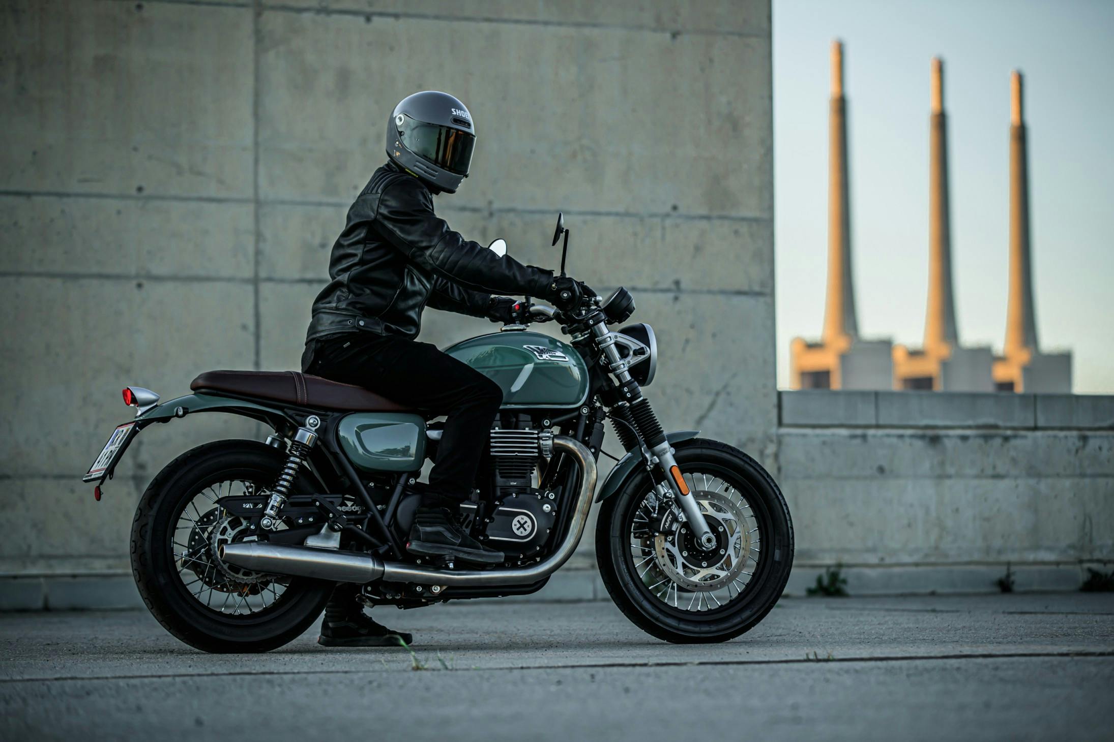 Rider in a leather jacket and a closed full helmet sitting on top of a Brixton Cromwell 1200 in Cargo Green in an industrial area
