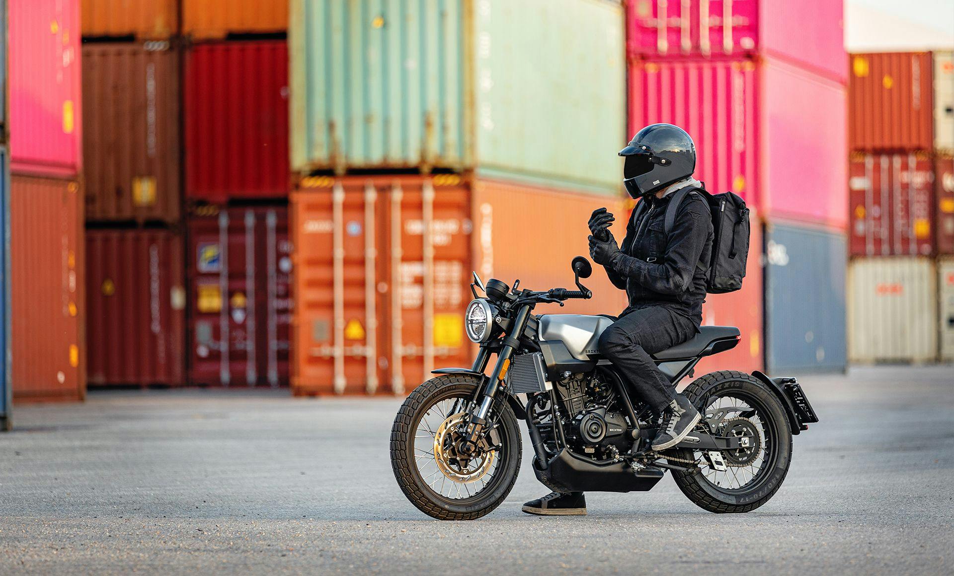 A landscape photo of motorcycle rider with a closed full helmet sitting on a Brixton Crossfire 125 in Bullet silver in front of  colourful shipping containers