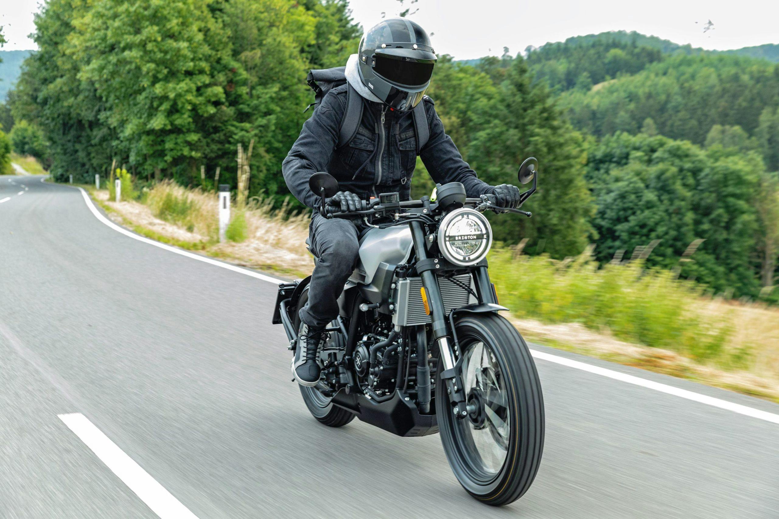 A rider wearing a closed full helmet and a Brixton backpack while riding a Brixton Crossfire 125 in Bullet Silver on a countryside road