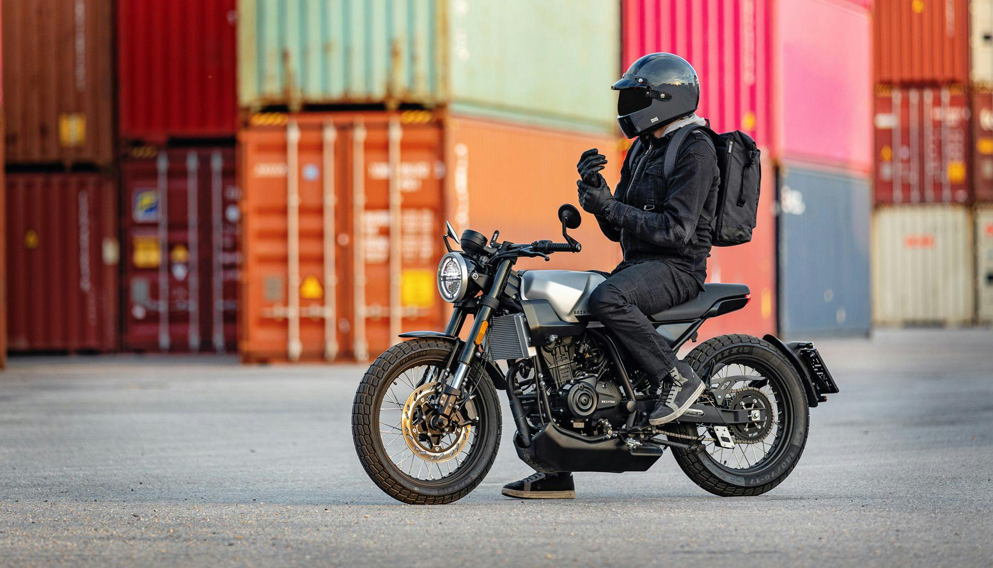 A landscape photo of motorcycle rider with a closed full helmet sitting on a Brixton Crossfire 125 in Bullet silver in front of colourful shipping containers