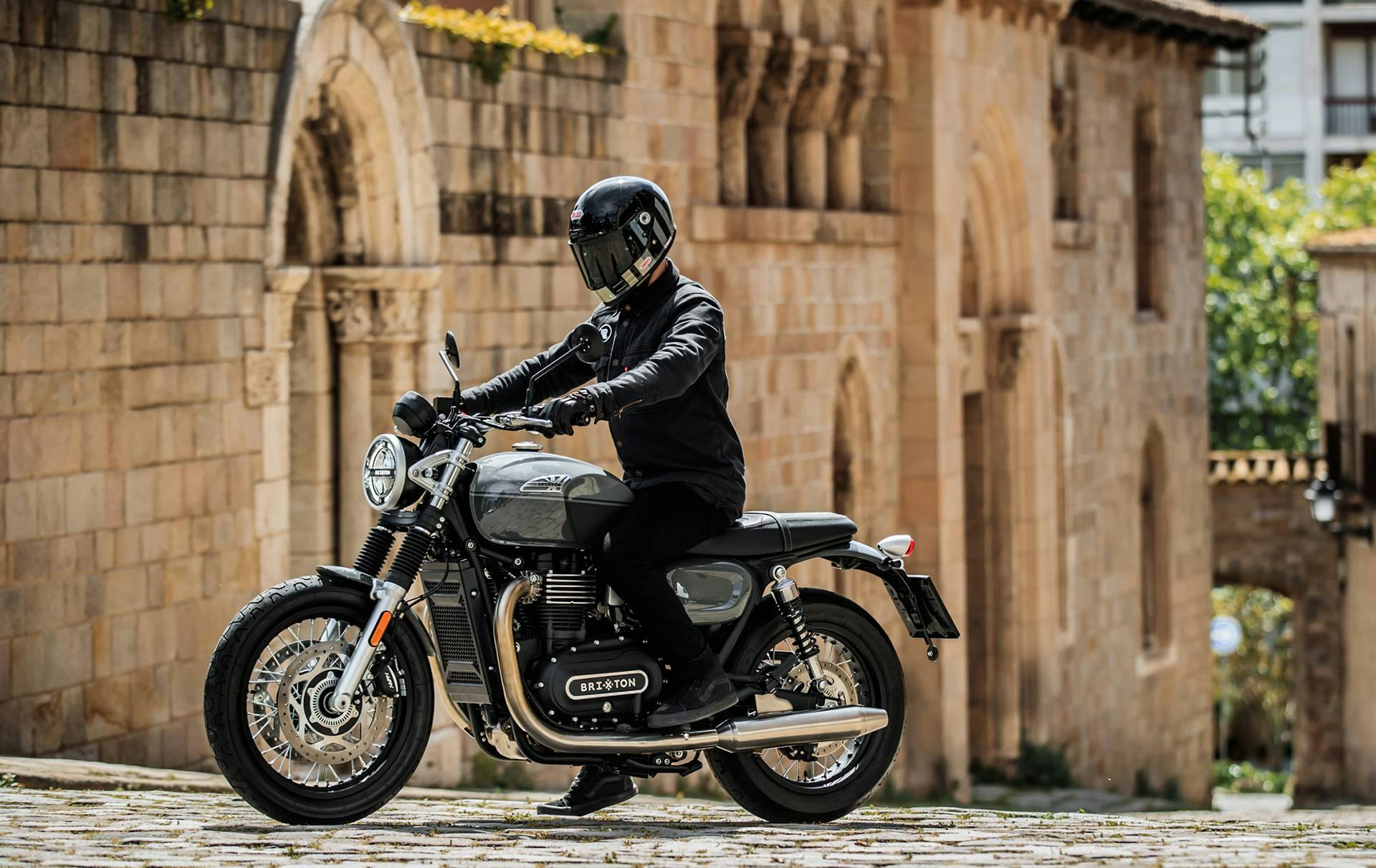 A motorcycle rider dressed in all black sitting on a Brixton Cromwell 1200 in Timberwolf Grey in front of an old building in Barcelona, Spain