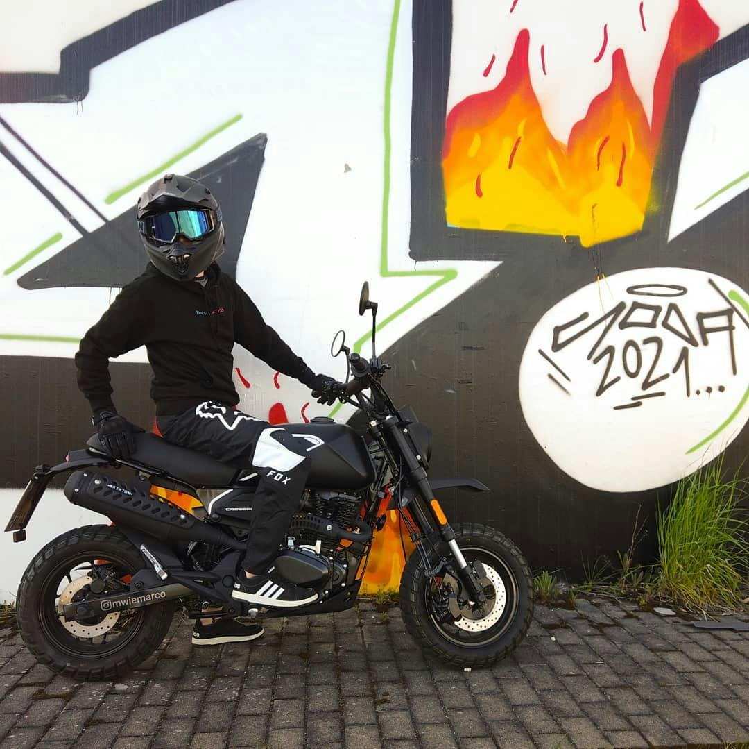 Rider of the Month Marco wearing his motorcycle helmet and googles while sitting on his Brixton Crossfire 125 XS in Backstage Black in front of a graffiti wall