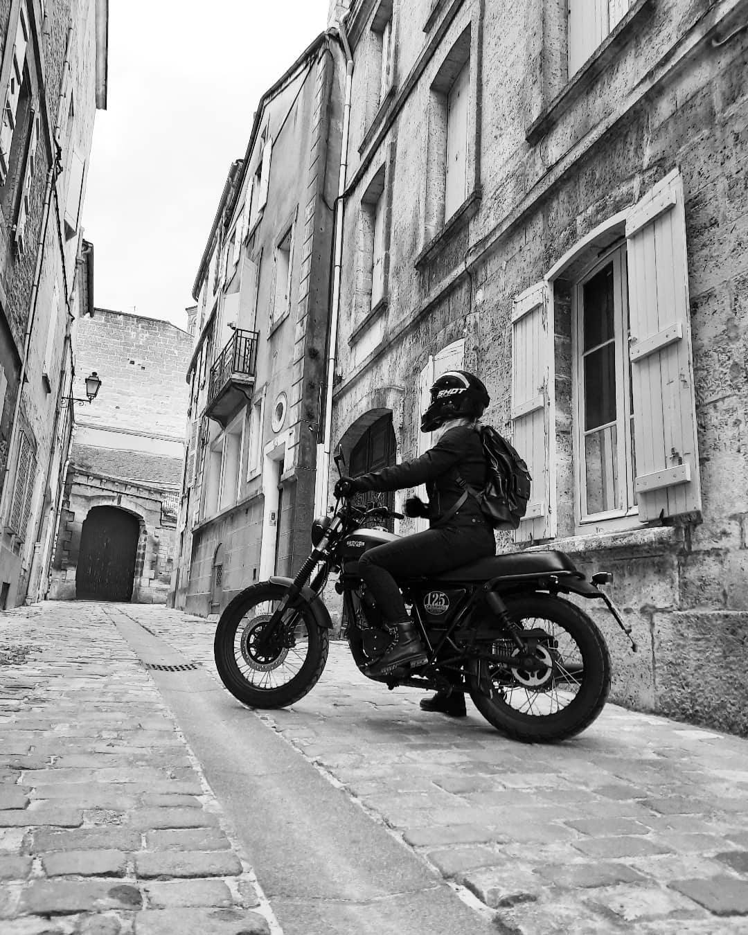 A black & white portrait photo of Rider of the Month Joss on her Cromwell 125 in Backstage Black on a cobbled old-town street