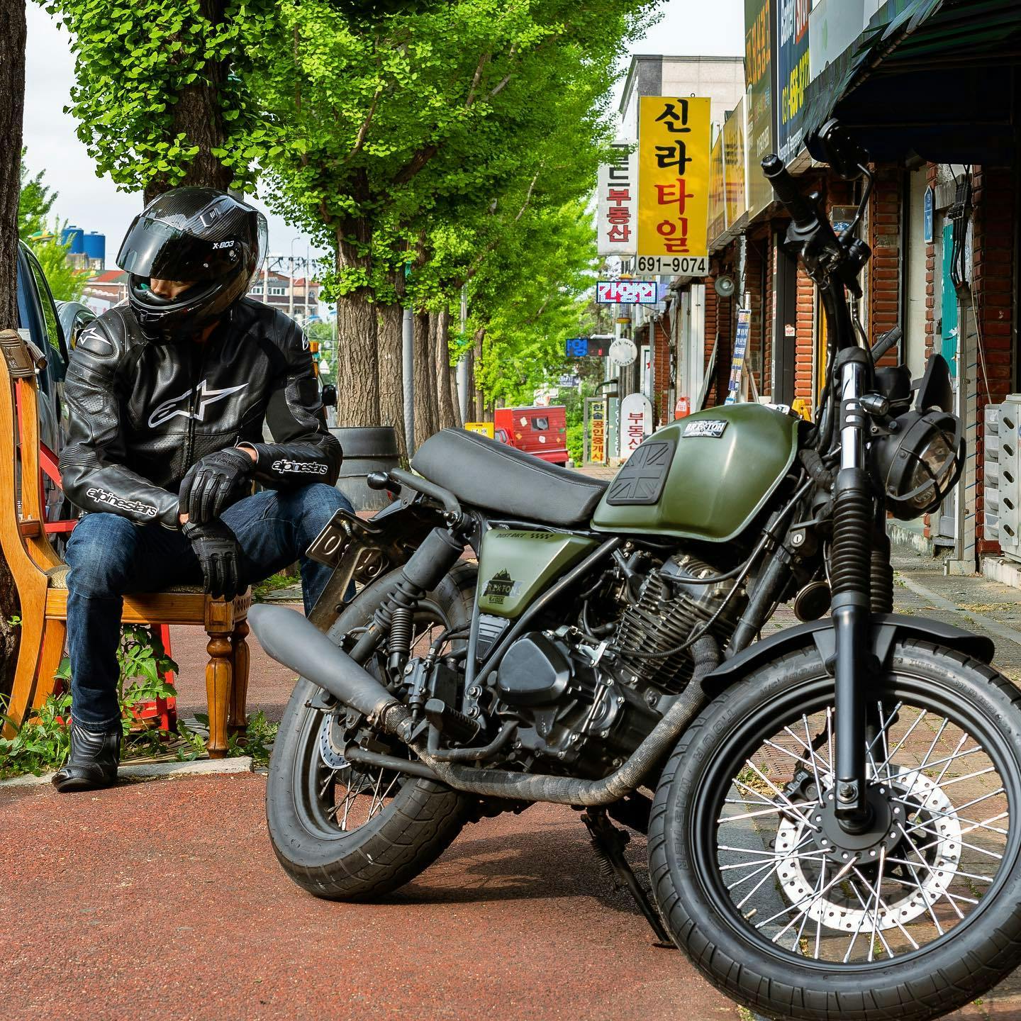 Brixton Felsberg 125 in Cargo Green parked on the sidewalk of an Asian country with the rider sitting in on a wooden chair in the background, the visor of his full helmet open