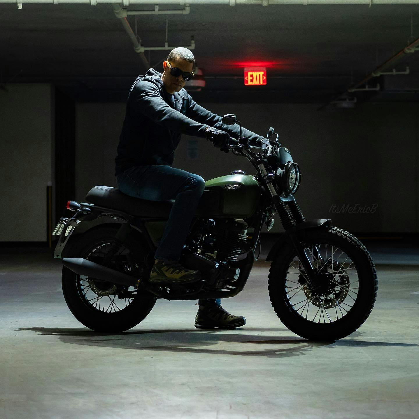 Brixton Felsberg 125 in Cargo Green standing in a dark garage with a rider sitting on top of it wearing a hooded vest and sunglasses, with an EXIT sign lit in the back