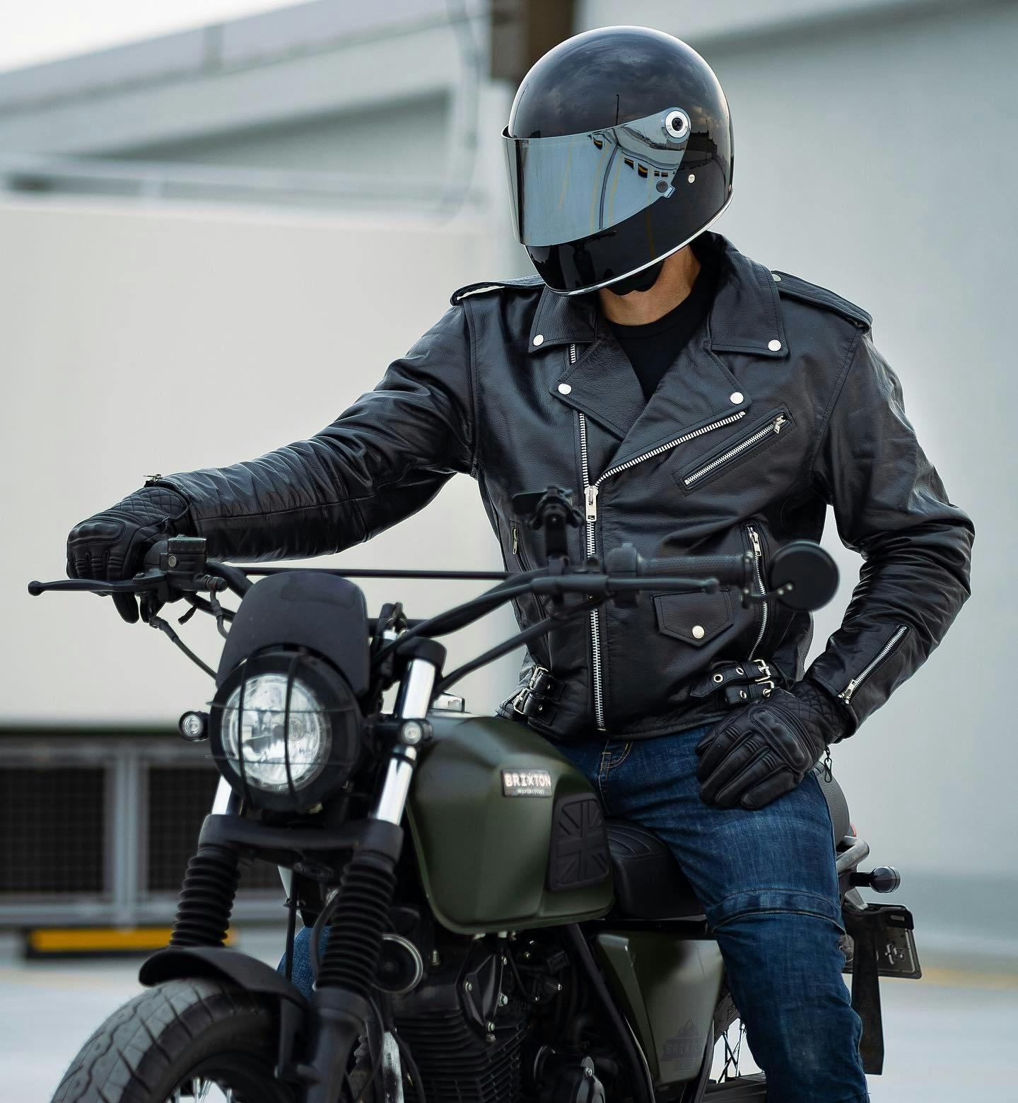 Portrait close-up of a Brixton Felsberg 125 in Cargo Green parked in front of a warehouse with a rider wearing a leather jacked and a full helmet with a closed visor sitting on top of it